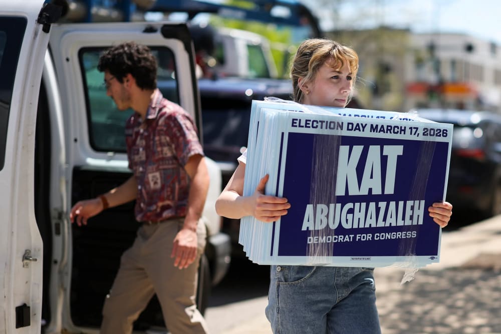 Kat Abughazaleh carries her signs on the sidewalk while a man walks towards a van in the background.
