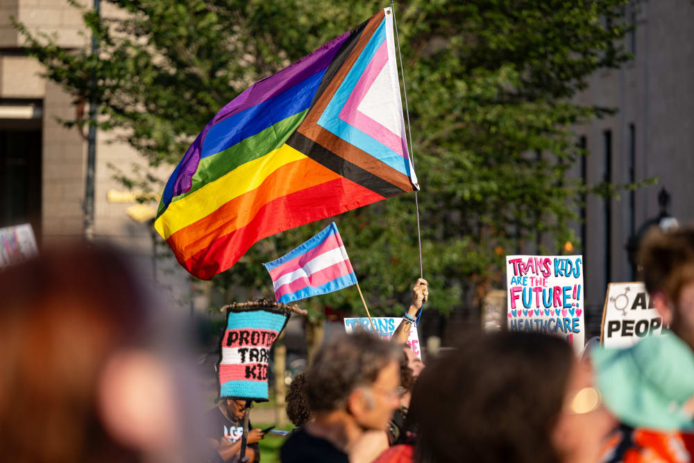 People hold signs in support of trans kids and LGBTQ+ flags.