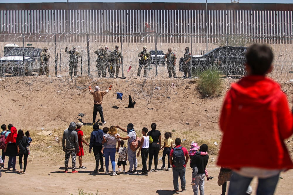 Adults and children with their backs to the camera look up at people in uniform who are behind a barbed wire fence.