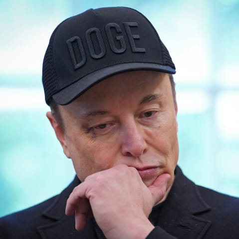 Elon Musk, wearing a black hat that says "DOGE," puts his hand to his chin while looking down.