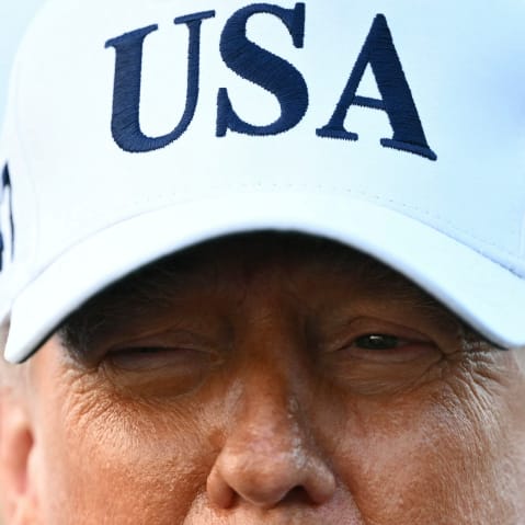 A closeup photo of President Trump shows the upper half of his face wearing a white USA hat.
