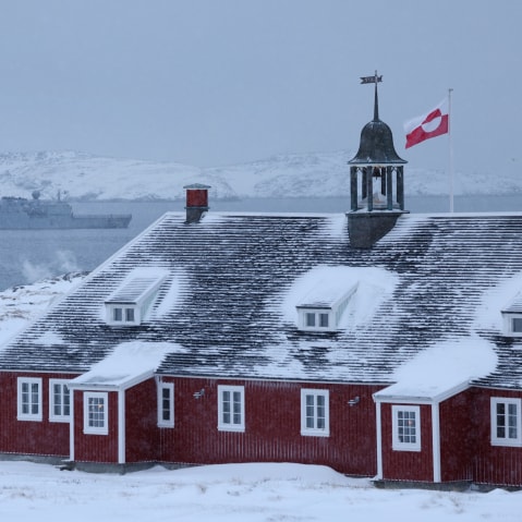 Greenland flag flies over building with Danish Navy ship in background