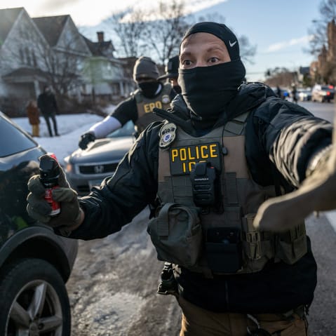 An ICE agent signals "stop" with his left hand up while holding a spray bottle as other agents follow him.