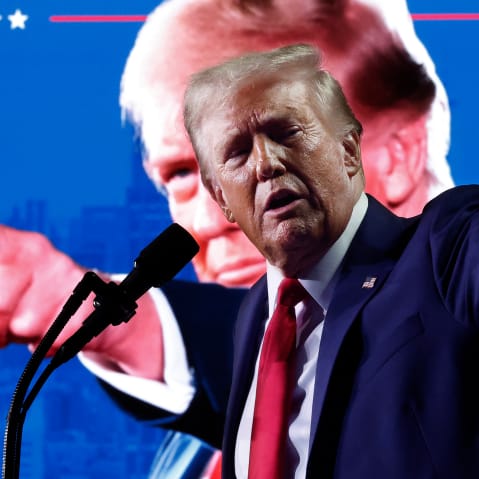 Donald Trump points at the crowd from the stage. His photo doing the same thing can be seen in the background.