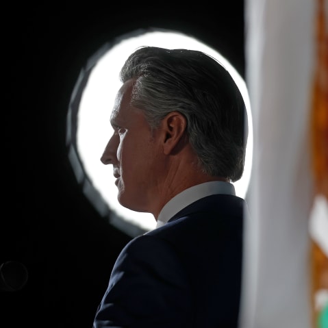 Gavin Newsom backlit, next to a California state flag.