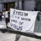 Two people hold a white blanket sign with black text that reads "Epstein is the worst kind of virus"