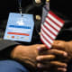A person seated, focus is on a badge they're wearing that reads "naturalization candidate" as they hold an American flag