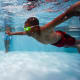 A child wearing goggles swims underwater in a pool, two other children are seen standing in the pool