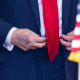 A closeup of Donald Trump's right hand is seen as he buttons his jacket behind an American flag.