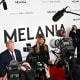 Donald and Melania Trump stand in front of a step-and-repeat banner advertising the movie "Melania." In front of them are journalists with cameras.