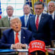 President Donald Trump, center, surrounded by members of the House and Senate in the Oval Office. Out of focus on the desk is a red hat that says "AMERICA IS BACK!"