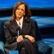 Kamala Harris sits at the stage while smiling and her hands are crossed over her legs.