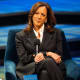 Kamala Harris sits at the stage while smiling and her hands are crossed over her legs.