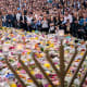 A crowd of people surrounds a large memorial filled with flowers. A menorah stands in the foreground.
