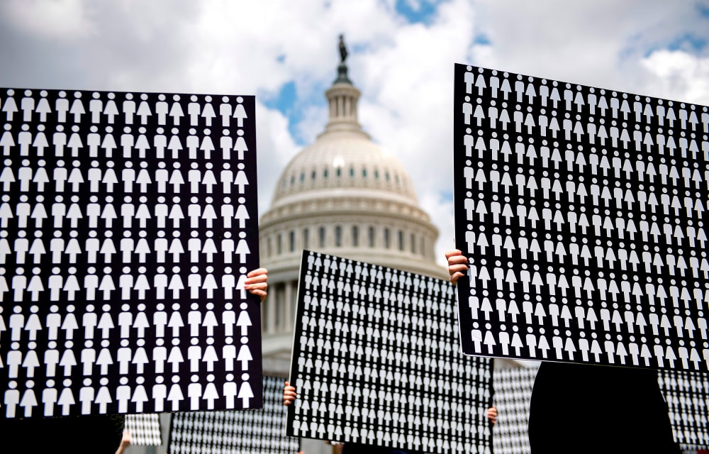 Image: Hands holding up placards representing people against the Capitol Hill in the background.