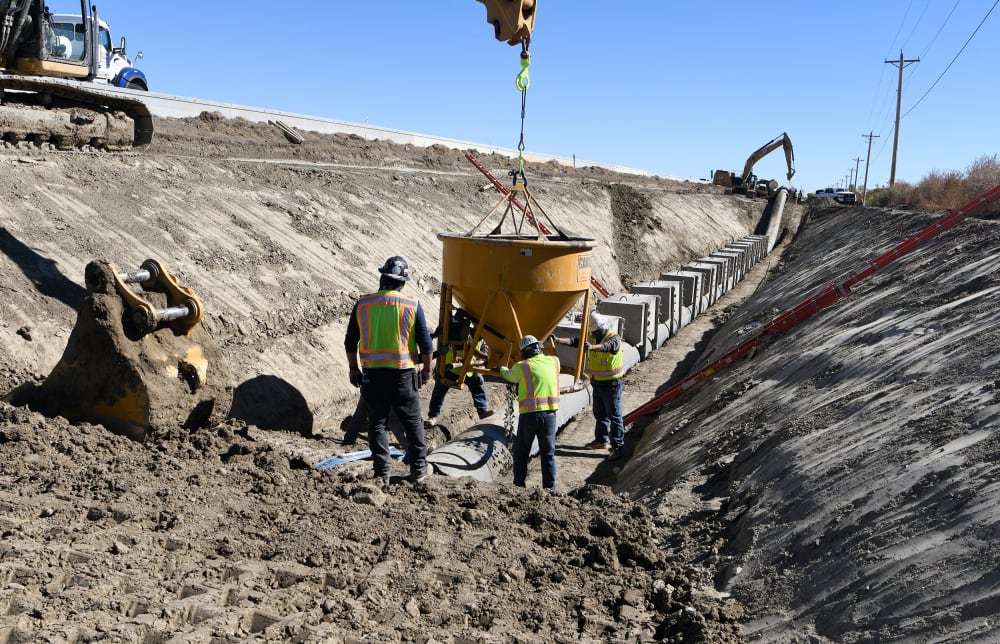 Workers in yellow high-visibility safety garments and hard hats set a concrete bucket on the ground of a construction zone.