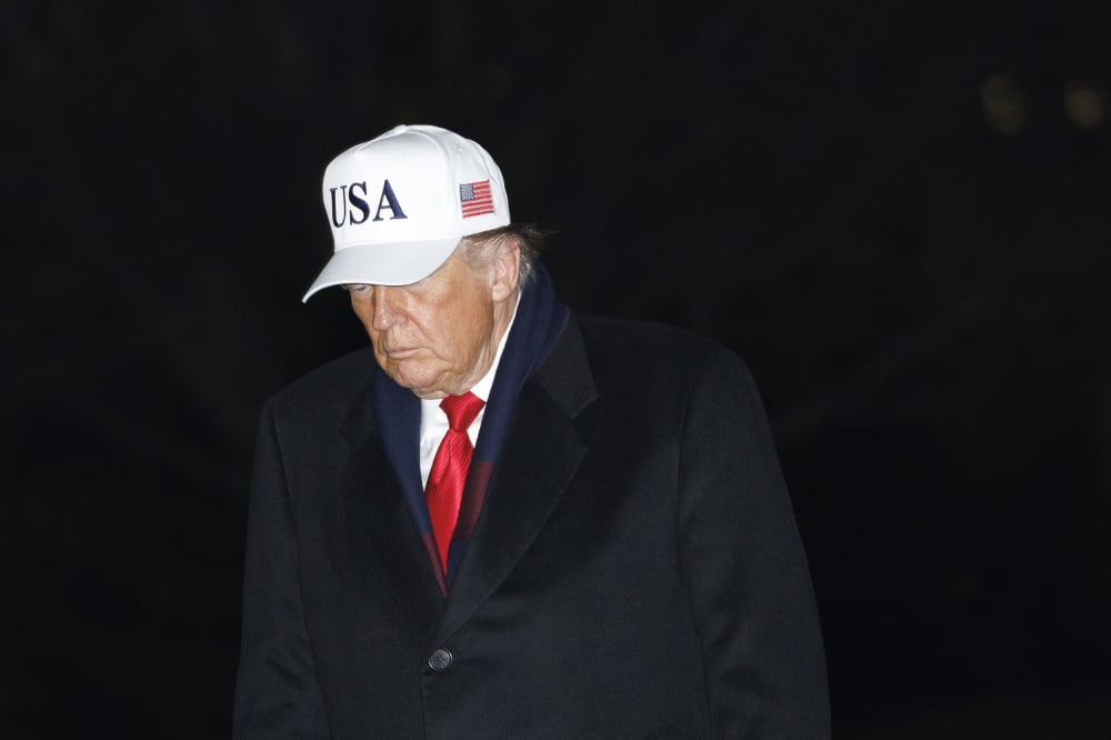 Donald Trump walks at the White House at night, wearing a white USA hat.