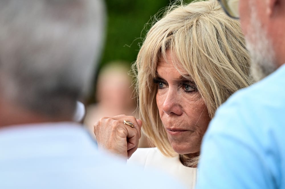 Brigitte Macron, right, looks on while standing with two males. This is a closeup frame.