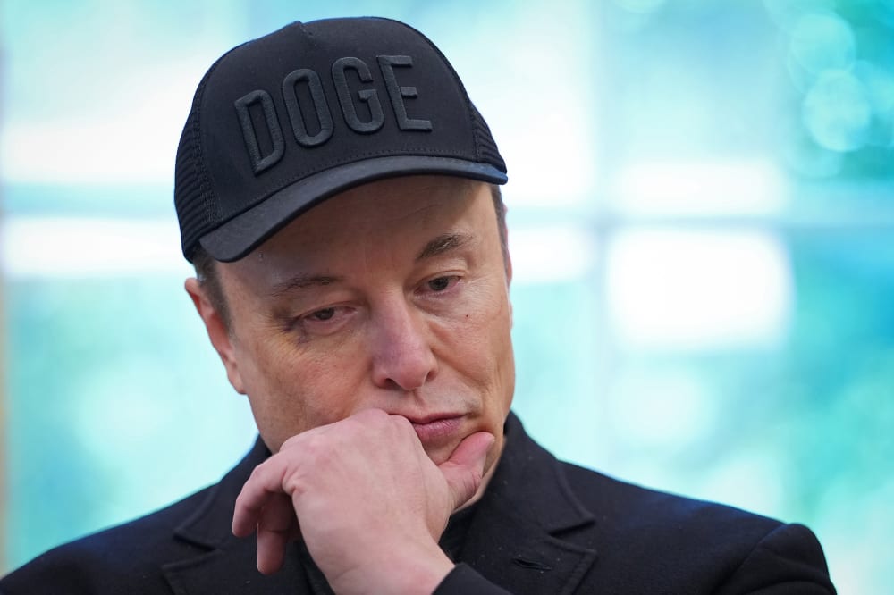 Elon Musk, wearing a black hat that says "DOGE," puts his hand to his chin while looking down.