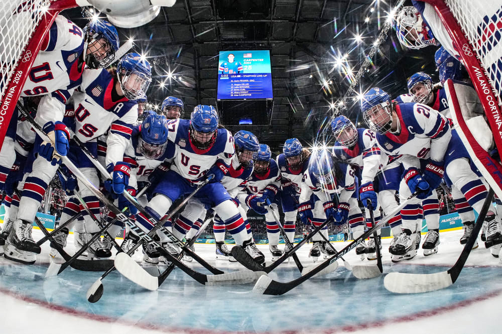 Hockey players in jerseys that read "USA" gather at the goal post. The photo is taken from a camera inside the goal.