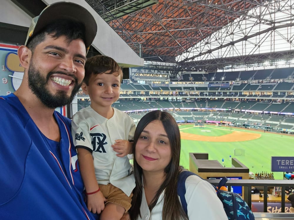 A man, woman, and small child, all in baseball jerseys. Behind them is a baseball field.