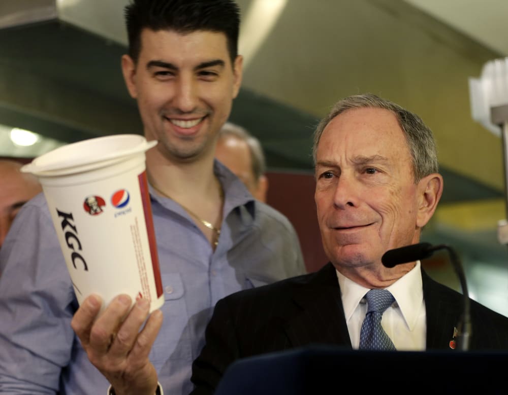 New York City Mayor Michael Bloomberg looks at a 64oz cup, as Lucky's Cafe owner Greg Anagnostopoulos, left, stands behind him, during a news conference at the cafe in New York, Tuesday, March 12, 2013. (AP Photo/Seth Wenig)