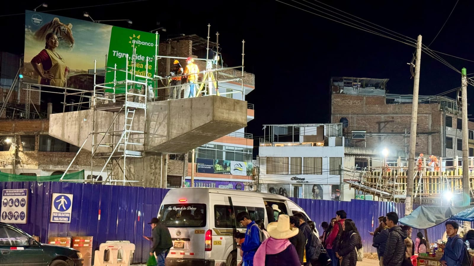 De noche, una obra de concreto a medio hacer sobre una avenida, con dos obreros con casco en lo alto de un andamio. Abajo, un cerco azul con un letrero de paso para peatones, una combi y gente caminando