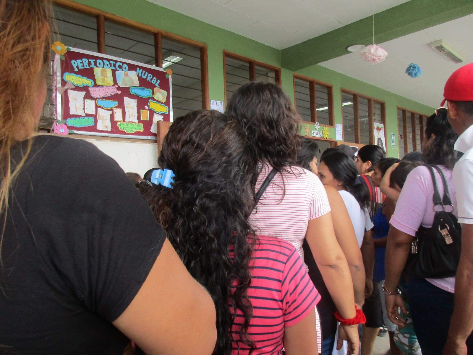 Una fila de personas, vistas de espaldas, en el pasadizo de un colegio, junto a un periódico mural y las ventanas de las aulas