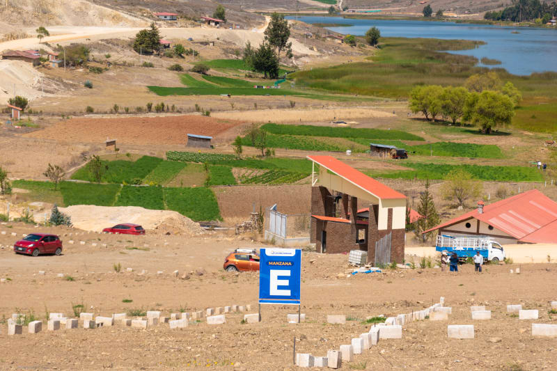 Lotes marcados con hitos de concreto y el cartel de la manzana E, con el pórtico de ingreso y la laguna al fondo entre chacras