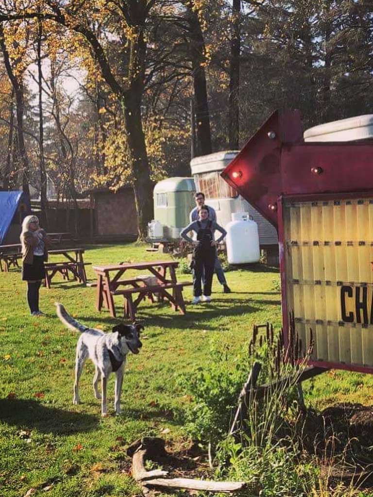 Group of people enjoying outdoor activities at Sou’wester Lodge, with cozy trailers in the background, a dog playing nearby, and picnic tables set up for relaxation and bonding.