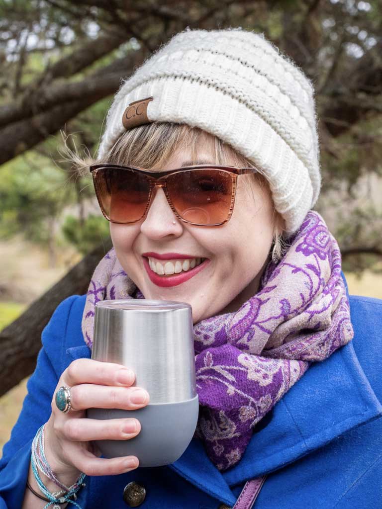 Smiling woman in sunglasses and a knit beanie, holding a stainless steel cup, wearing a blue coat and patterned scarf, enjoying a cozy outdoor retreat at Sou’wester Lodge in Seaview, Washington.