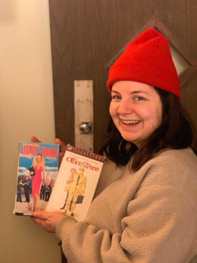 Person in a red beanie smiling while holding VHS tapes of "Legally Blonde" and "Office Space" in a cozy lodge setting, reflecting the fun movie night at Sou’wester Lodge.