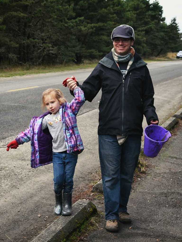 Adult man holding the hand of a young girl in a colorful jacket, walking along a road with a purple bucket, surrounded by trees, embodying a family-friendly outdoor activity during a retreat at Sou’wester Lodge.