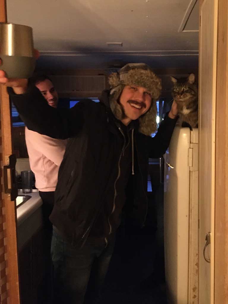 Man in a furry hat holding a drink, smiling in a cozy trailer kitchen, with a cat perched on the fridge, capturing a relaxed moment during a Murmur Creative retreat.