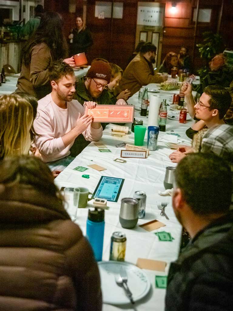Group of people engaged in a lively game night at Sou’wester Lodge, sharing laughter and drinks, with a focus on a player holding a game board, reflecting Murmur Creative's team-building retreat activities.