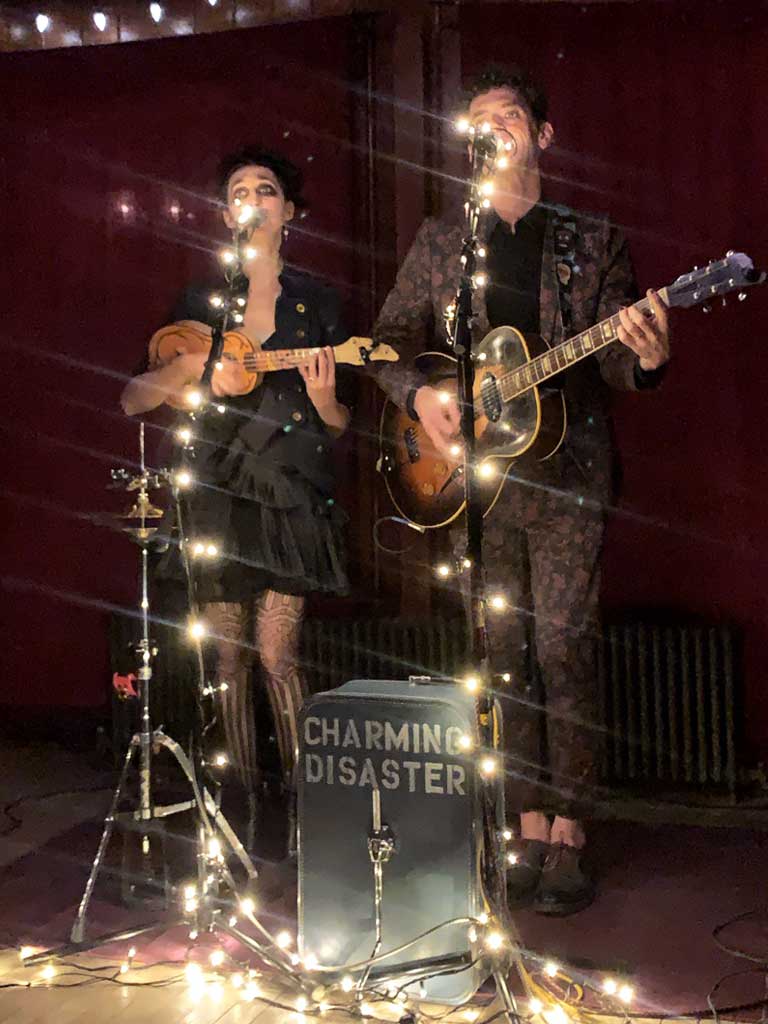 Musical performance by Charming Disaster, featuring a woman with a ukulele and a man with an acoustic guitar, illuminated by string lights in a cozy lodge setting.