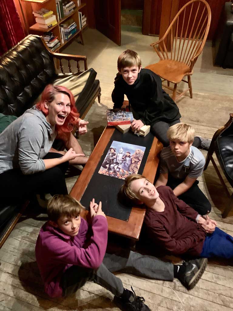 Group of children and an adult woman gathered around a table, excitedly pointing at a board game, in a cozy lodge setting with bookshelves and wooden decor, reflecting a fun bonding experience during a retreat at Sou’wester Lodge.