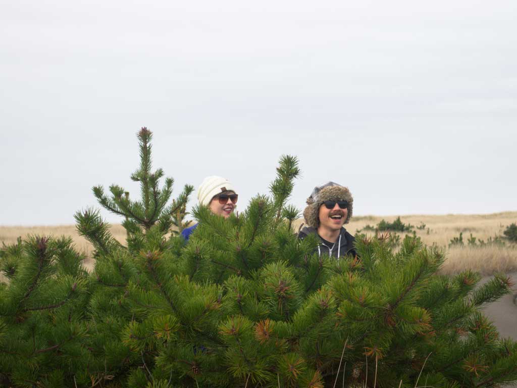 Two individuals smiling and peeking from behind a green pine tree, wearing winter hats and sunglasses, in a coastal outdoor setting during a Murmur Creative retreat.