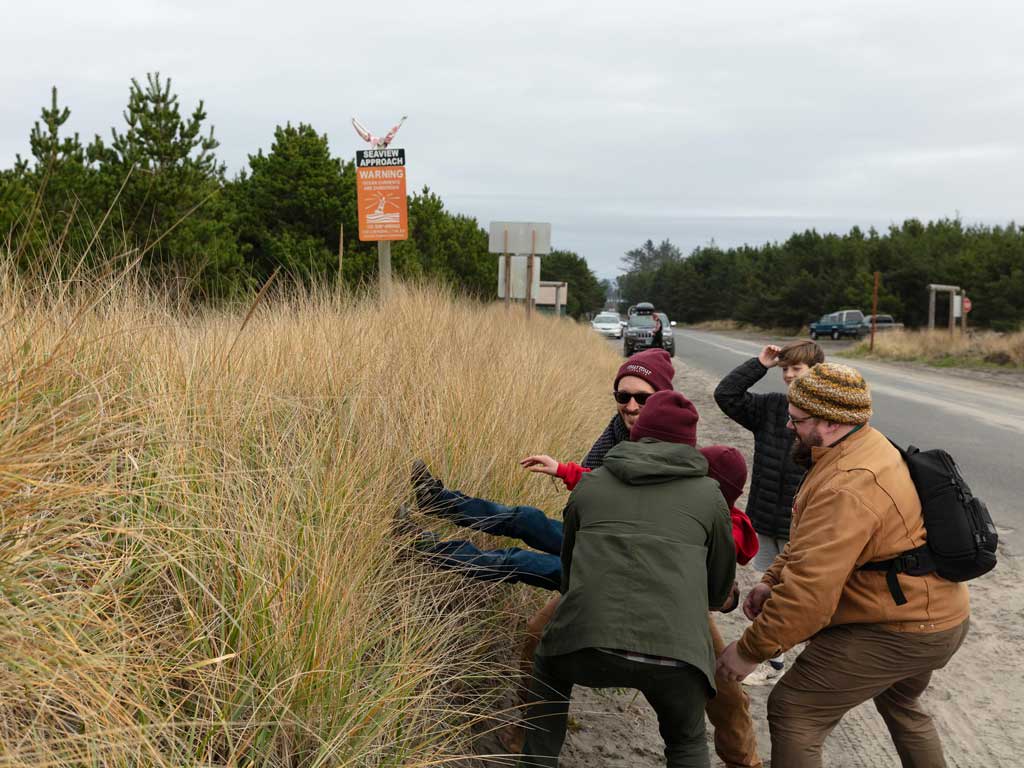 Group of friends laughing and engaging in a playful moment near the beach, surrounded by tall grass and a warning sign for Seaview, capturing a fun retreat experience at the Sou'wester Lodge.