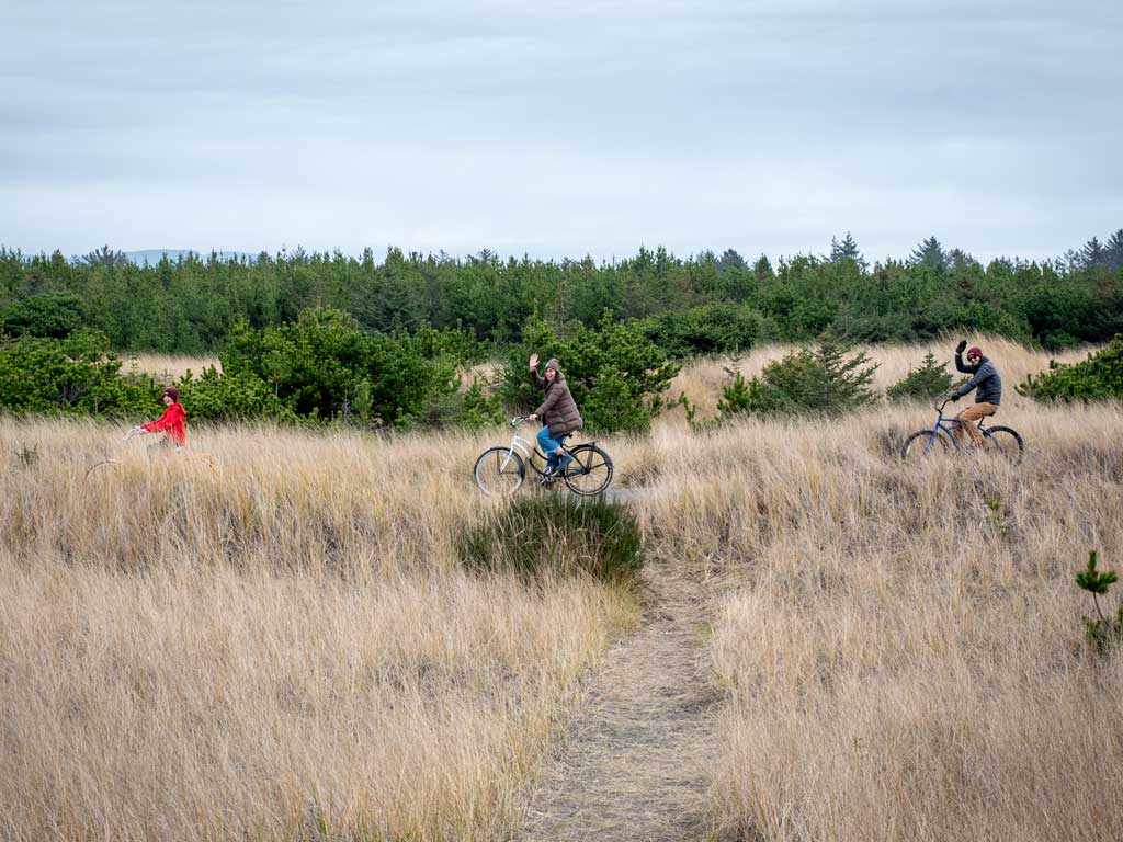 Three individuals biking through tall grass in a natural setting, enjoying outdoor activities during a retreat at Sou'wester Lodge, Seaview, Washington.