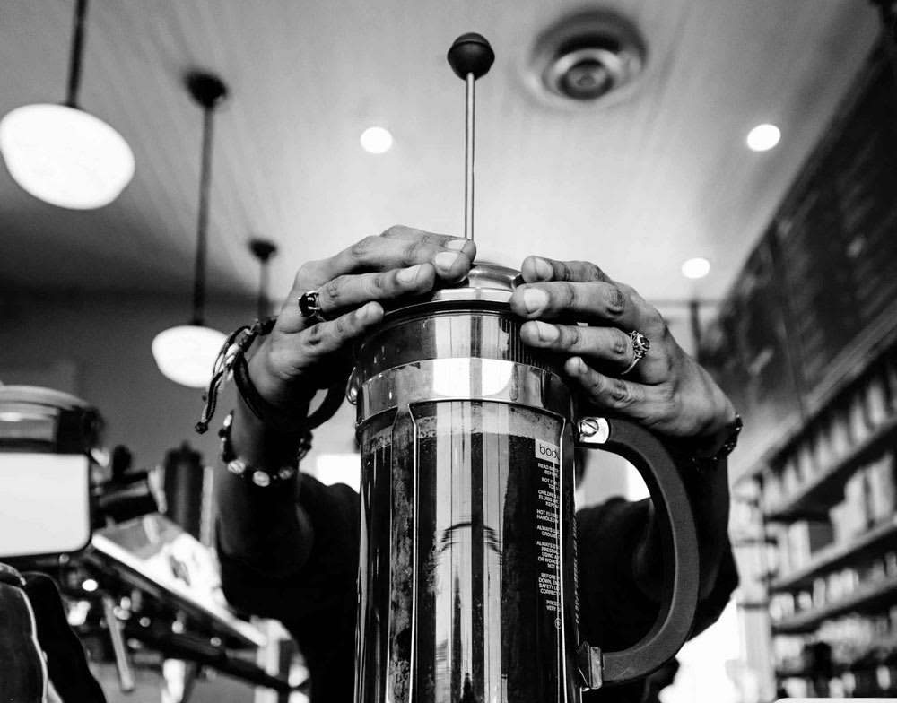 Hands pressing down on a French press coffee maker in a café setting, emphasizing the brewing process for coffee enthusiasts in the food and beverage industry.