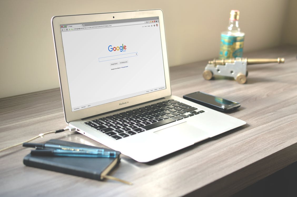 Laptop displaying Google search page, with a smartphone, notebook, and pen on a wooden desk, emphasizing online privacy concerns related to internet usage.
