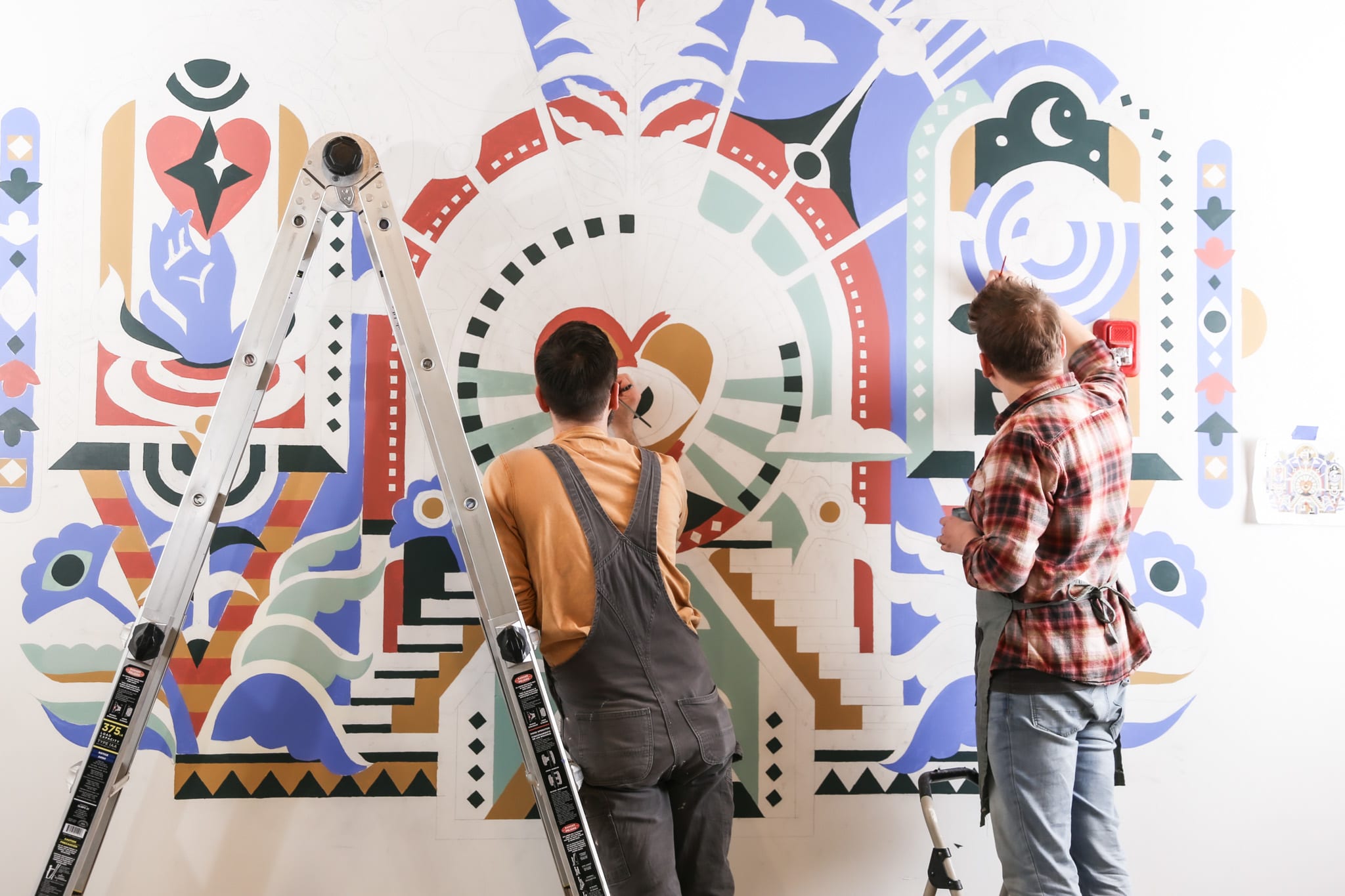 Two artists painting a colorful mural on a wall, featuring abstract shapes and designs, with a ladder nearby, reflecting collaboration and creativity at Murmur Creative in Portland.