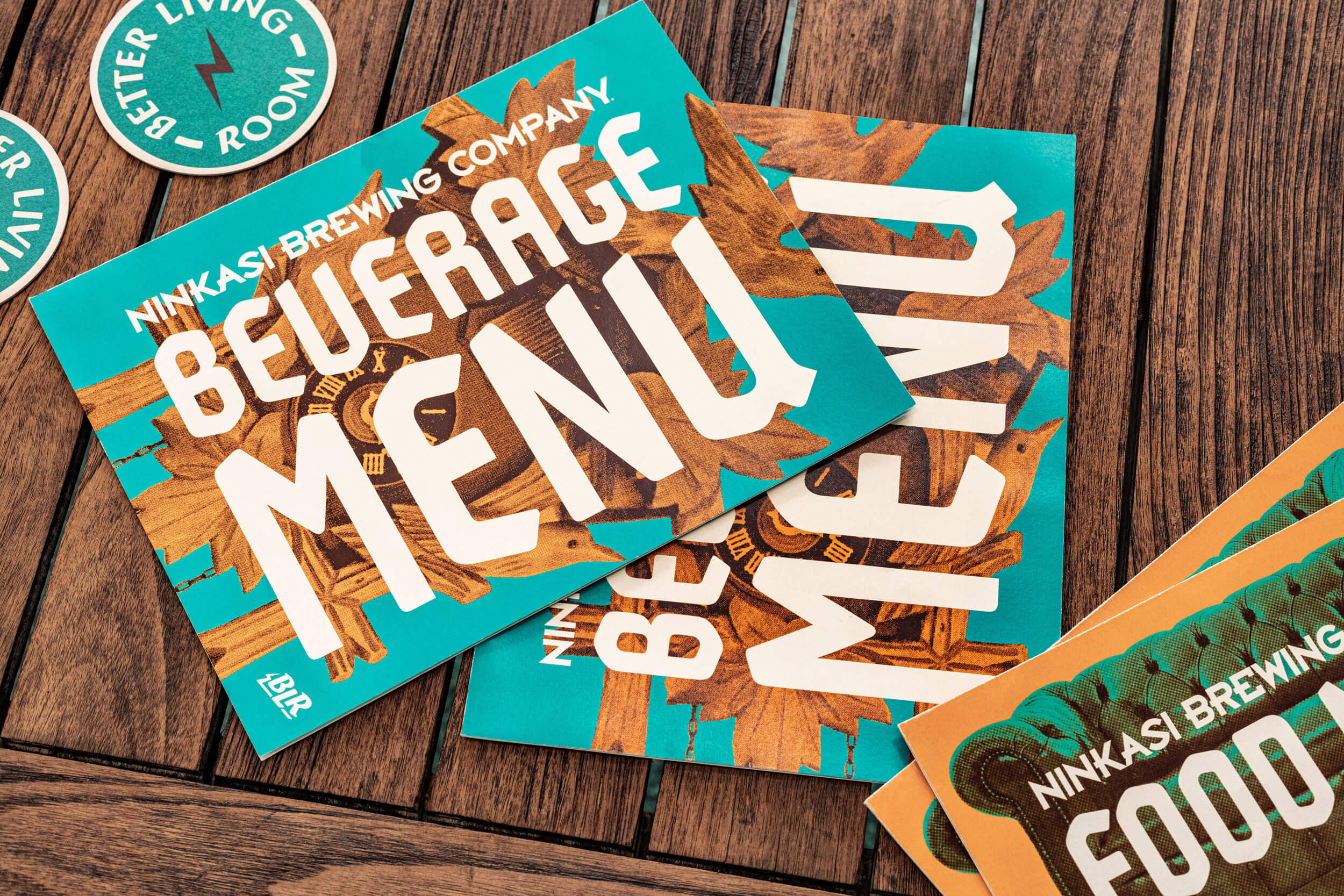 Ninkasi Brewing Company beverage menu showcasing a vintage design, featuring bold typography and colorful graphics, placed on a wooden table alongside coasters, reflecting the inviting atmosphere of the Better Living Room in Eugene, Oregon.