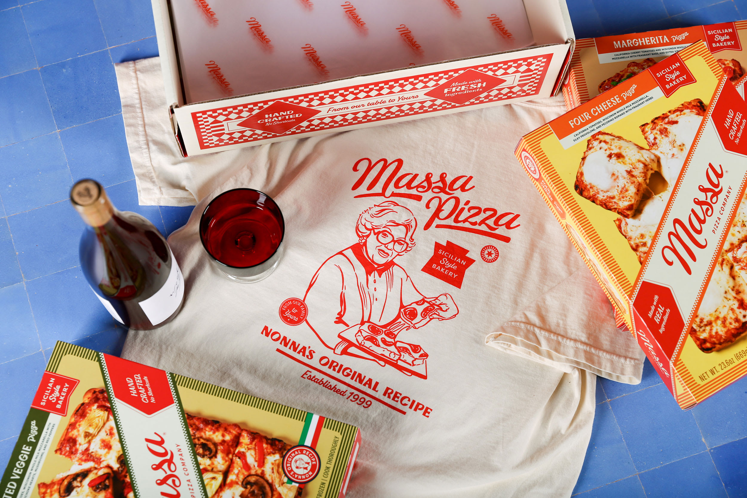 Massa Pizza branding elements including a vintage t-shirt with a Sicilian woman, pizza boxes for Margherita and Four Cheese varieties, a wine bottle, and a glass on a blue tiled surface, highlighting the brand's Italian heritage and modern packaging design.