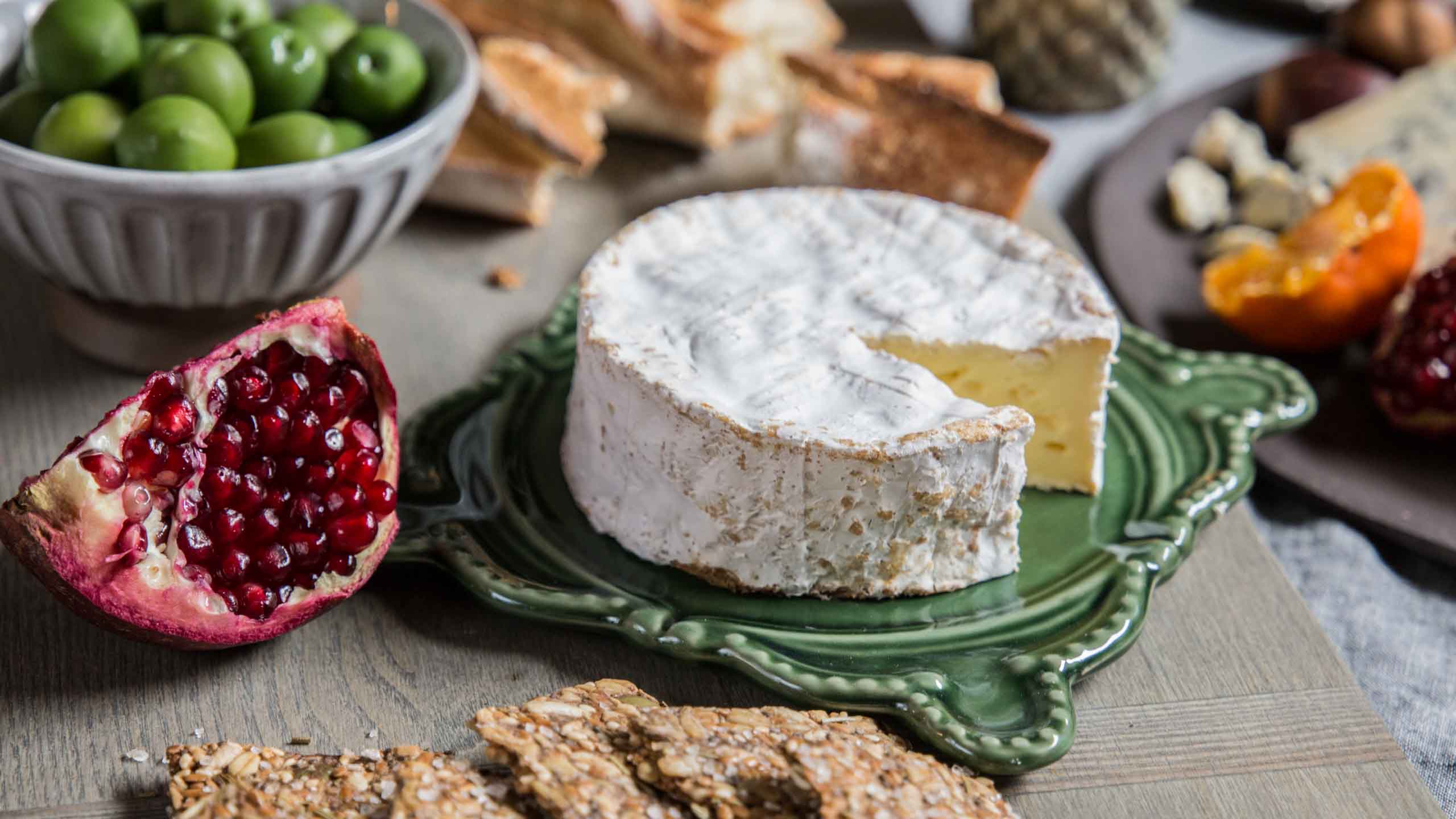 Cheese wheel on decorative green plate with pomegranate, green olives, and crackers, showcasing gourmet food arrangement for Zupan's Markets upscale customer experience.