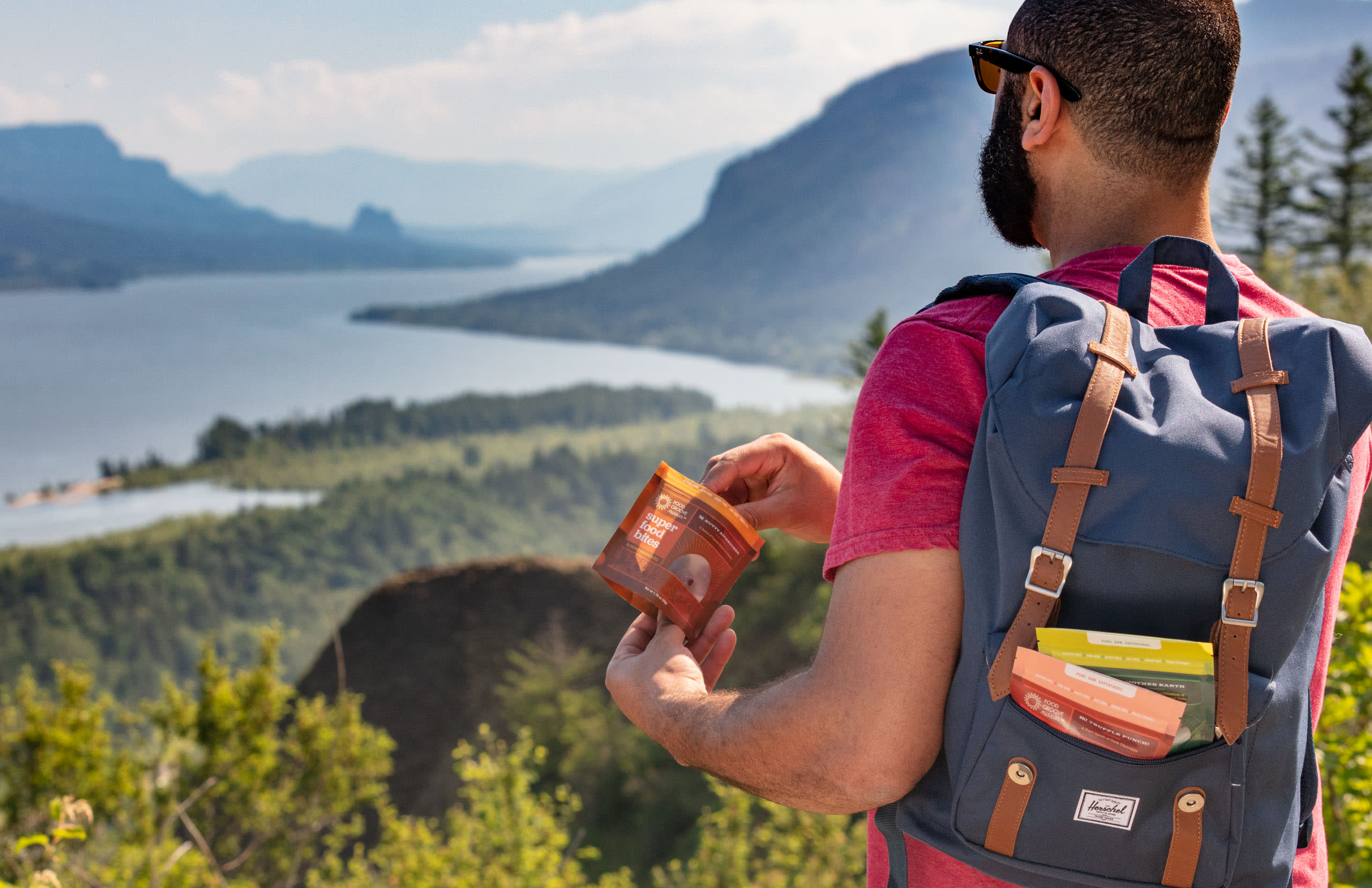 Person holding Food Groove Mission superfood bites packaging, with a scenic landscape of mountains and a river in the background, showcasing healthy snack options for outdoor enthusiasts.