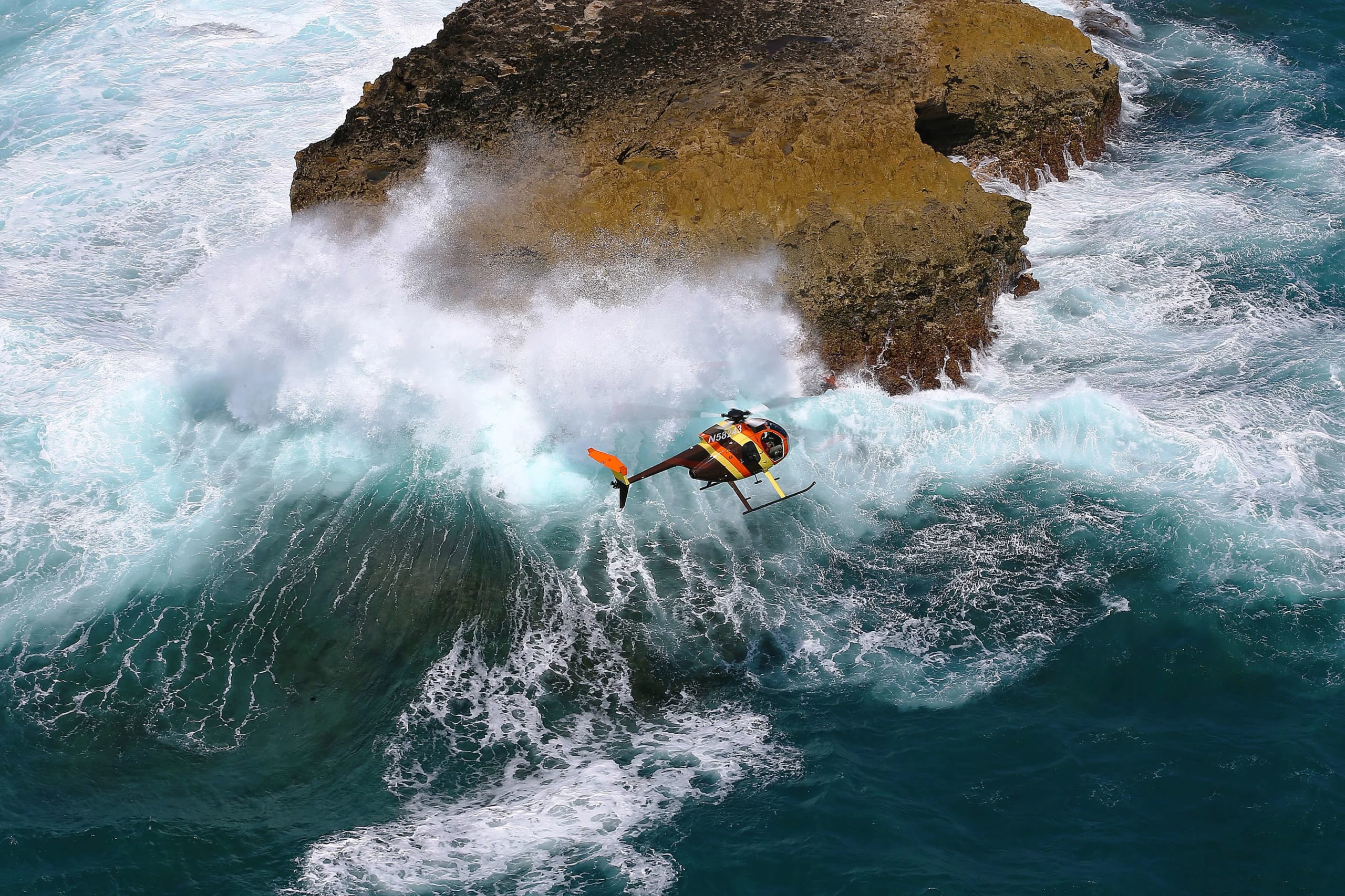 Helicopter flying over crashing waves near rocky coastline, showcasing Hawaii's stunning natural beauty and adventure experiences offered by Paradise Helicopters.