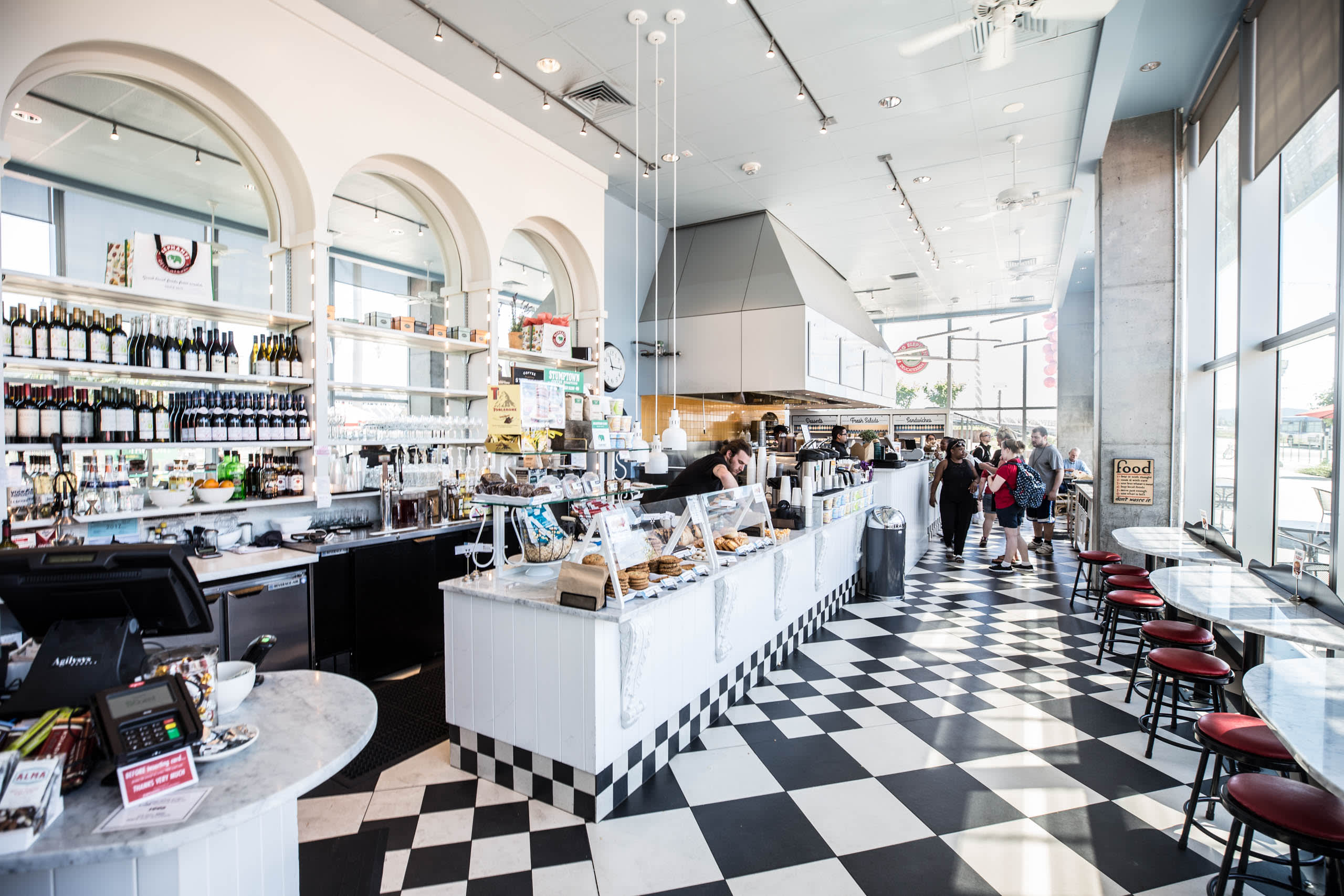 Brightly lit interior of Elephants Delicatessen showcasing a stylish café with a marble counter, wine shelves, and customers interacting, emphasizing the whimsical and inviting atmosphere of the gourmet food provider.