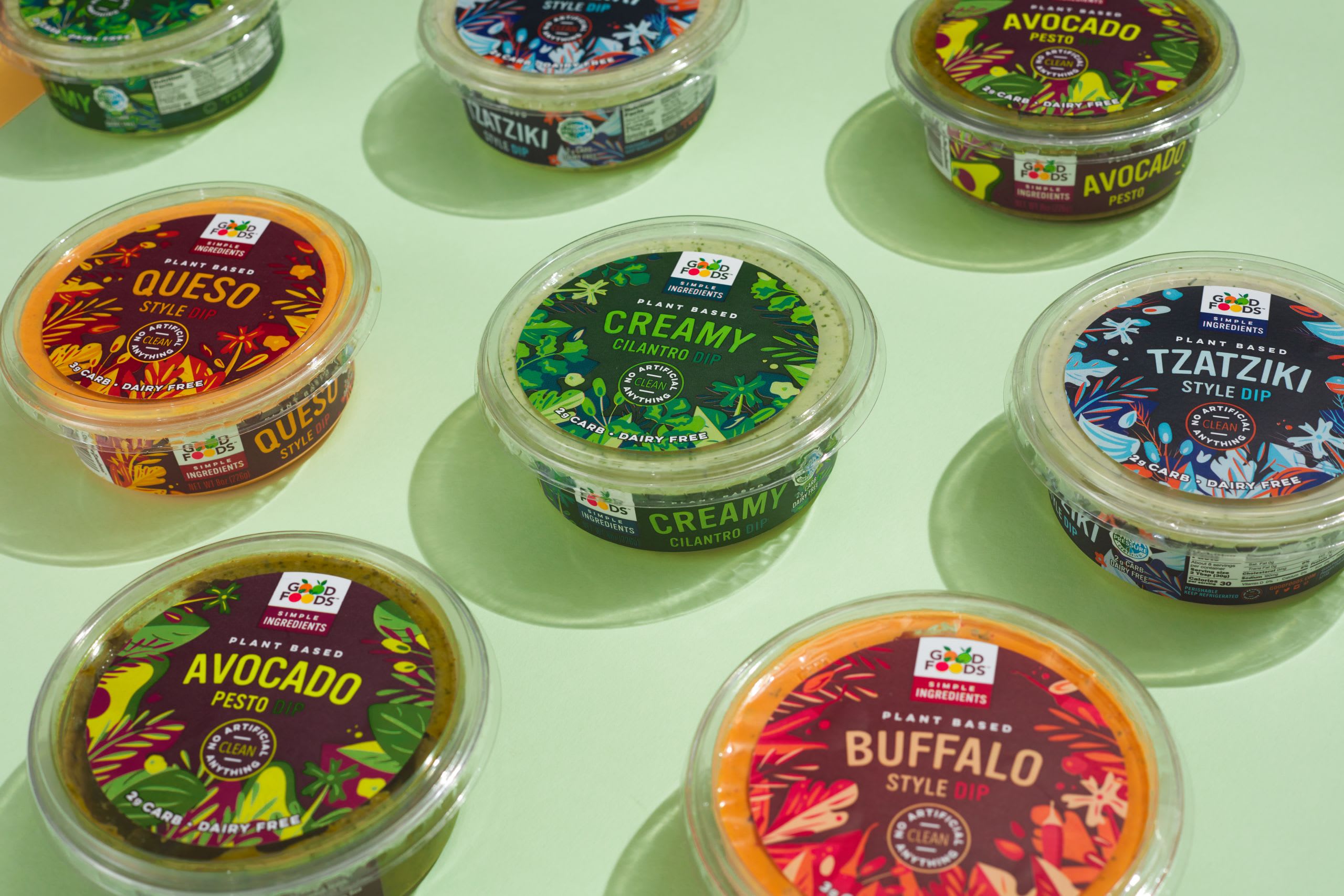 Colorful packaging of Good Foods' plant-based dips, featuring flavors like Queso, Creamy Cilantro, Avocado Pesto, and Tzatziki, on a light green background, emphasizing fresh ingredients and clean labels.